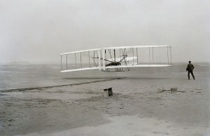 First successful flight of the Wright Flyer, by the Wright brothers. The machine traveled 120 ft (36.6 m) in 12 seconds at 10:35 a.m. at Kitty Hawk, North Carolina. Orville Wright was at the controls of the machine, lying prone on the lower wing with his hips in the cradle which operated the wing-warping mechanism. Wilbur Wright ran alongside to balance the machine, and just released his hold on the forward upright of the right wing in the photo. The starting rail, the wing-rest, a coil box, and other items needed for flight preparation are visible behind the machine. This was considered "the first sustained and controlled heavier-than-air, powered flight" by the Fédération Aéronautique Internationale.  Via Wikipedia