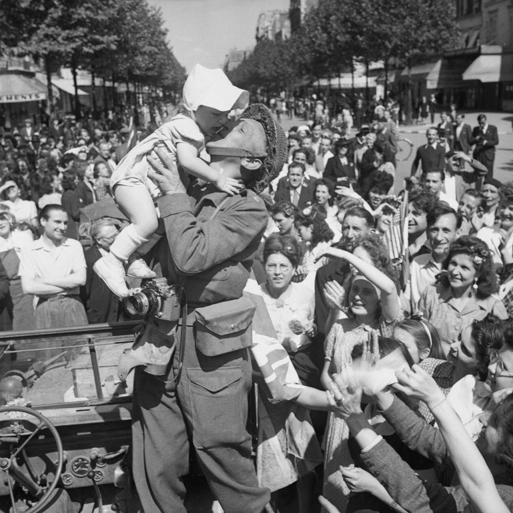 An AFPU photographer kisses a small child before cheering crowds in Paris, one day after liberation, 26 August 1944.